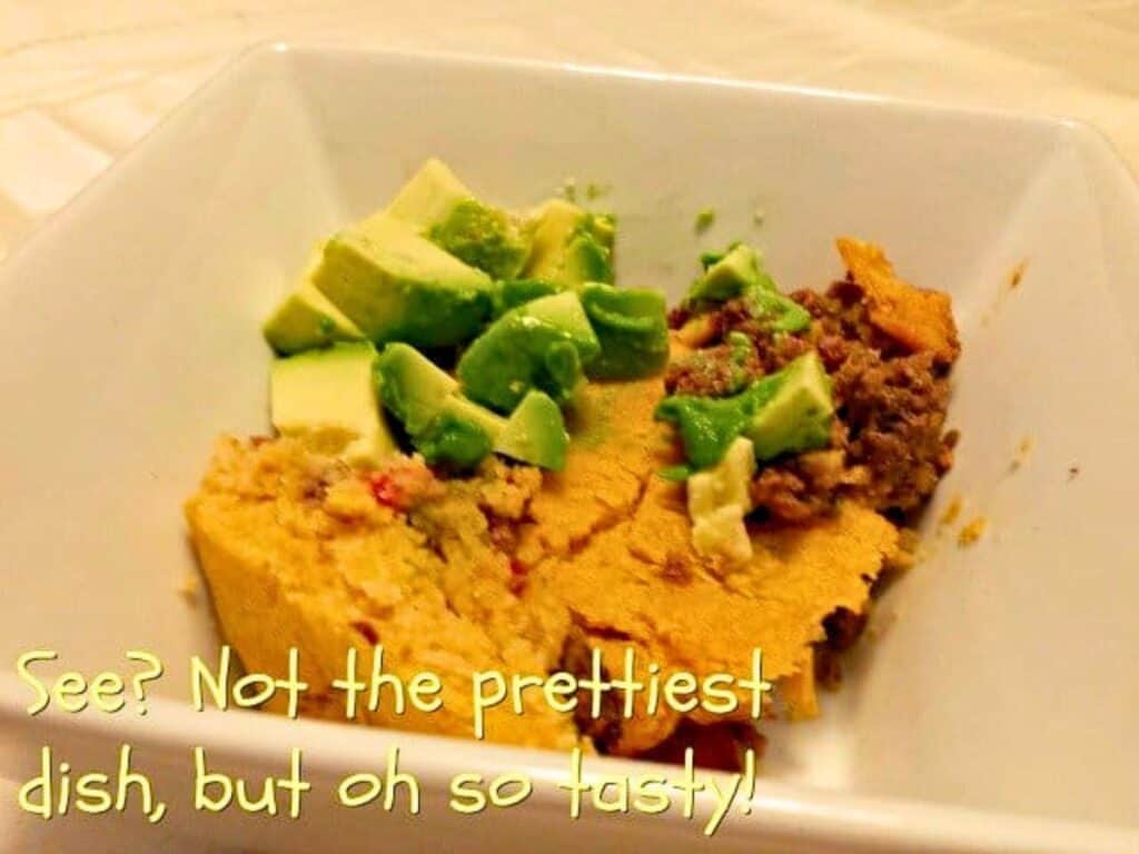 A square white bowl contains a serving of casserole topped with chopped avocado, ground meat, and cornbread. Yellow text at the bottom reads, &ldquo;See? Not the prettiest dish, but oh so tasty!&rdquo;.