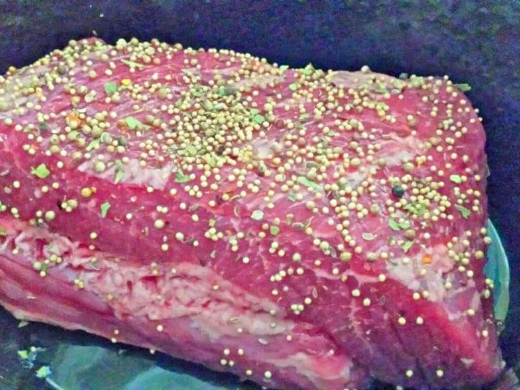 Raw corned beef brisket covered with pickling spices, resting in a dark cooking pot.