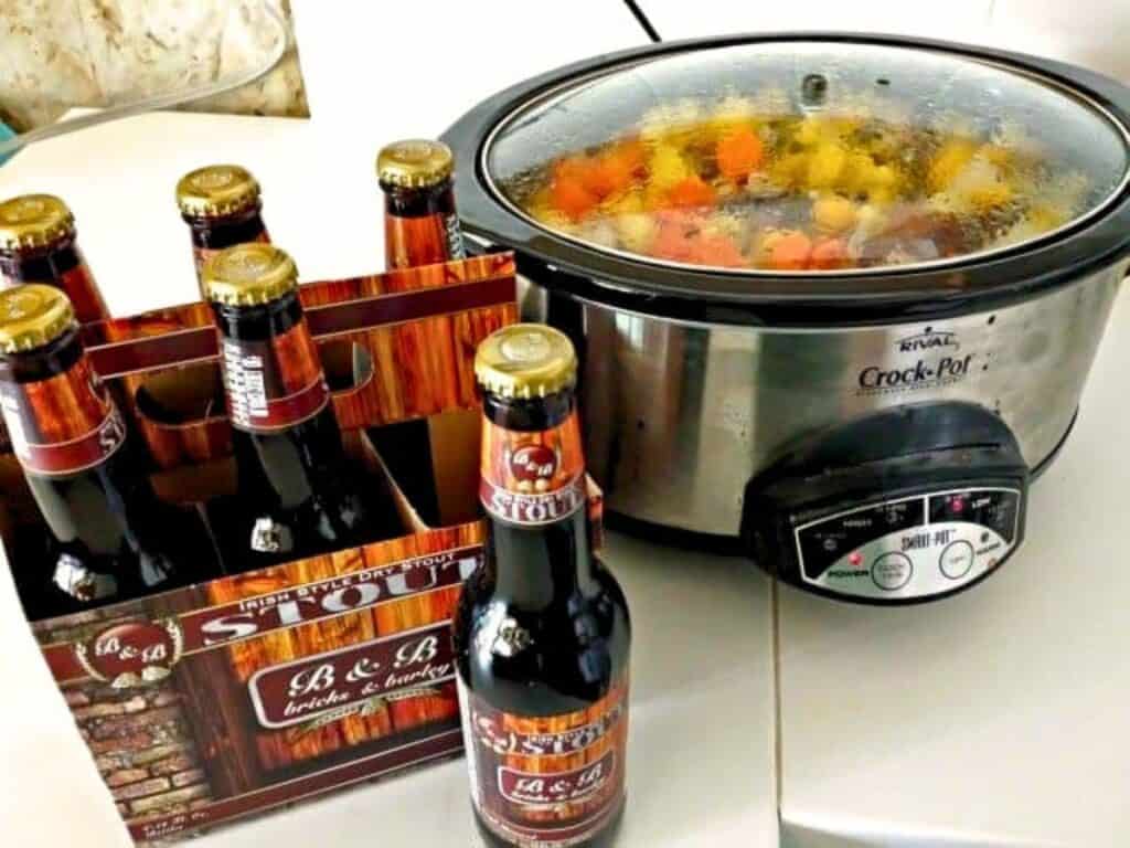 A slow cooker with stew inside sits next to a six-pack of bottled stout beer on a white countertop.