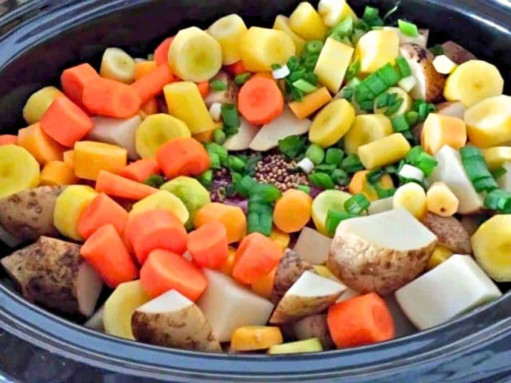 Chopped carrots, potatoes, parsnips, and green onions are arranged in a slow cooker with visible mustard seeds in the center.