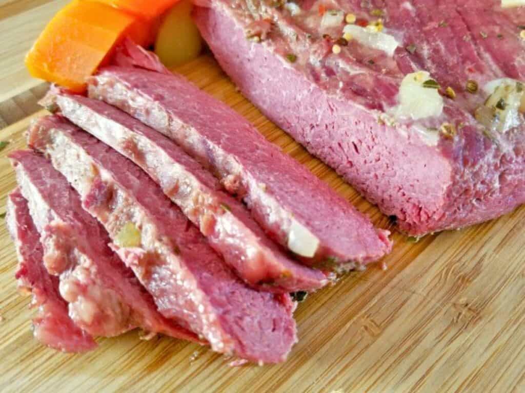 Sliced corned beef with visible fat and spices on a wooden cutting board, next to pieces of carrot.