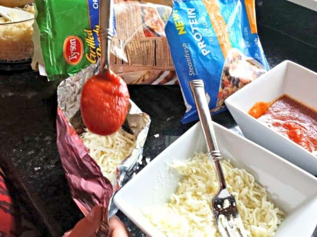 A person spoons tomato sauce over shredded cheese next to a bowl of cheese, a bowl of sauce, and packaged chicken and vegetables on a kitchen counter while preparing grilled chicken pockets.