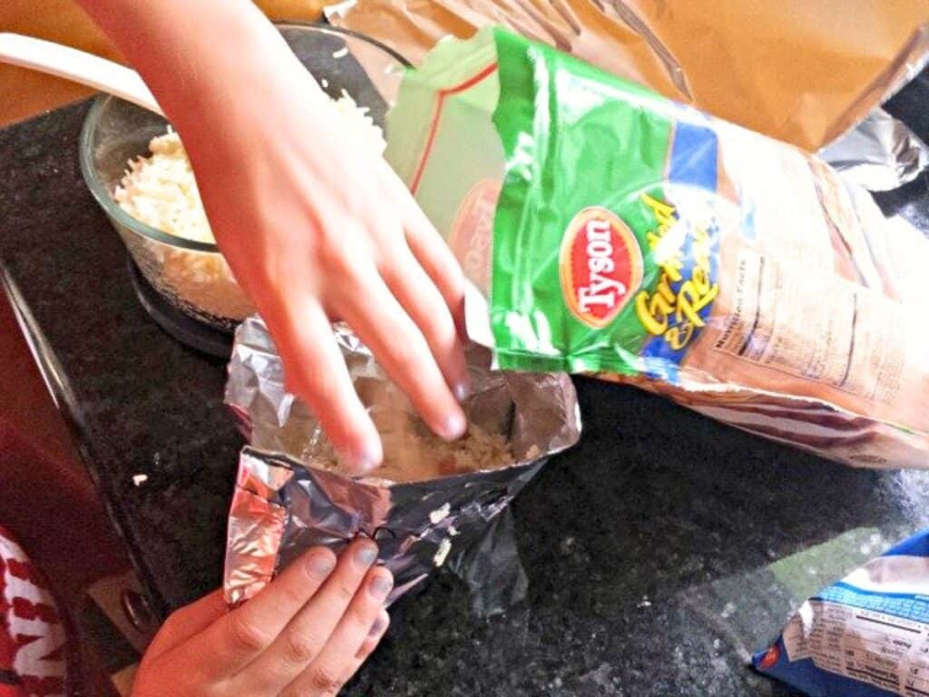 Two hands reach into an open bag of Tyson grilled chicken on a black countertop next to a bowl of shredded cheese, ready to create delicious grilled chicken pockets.