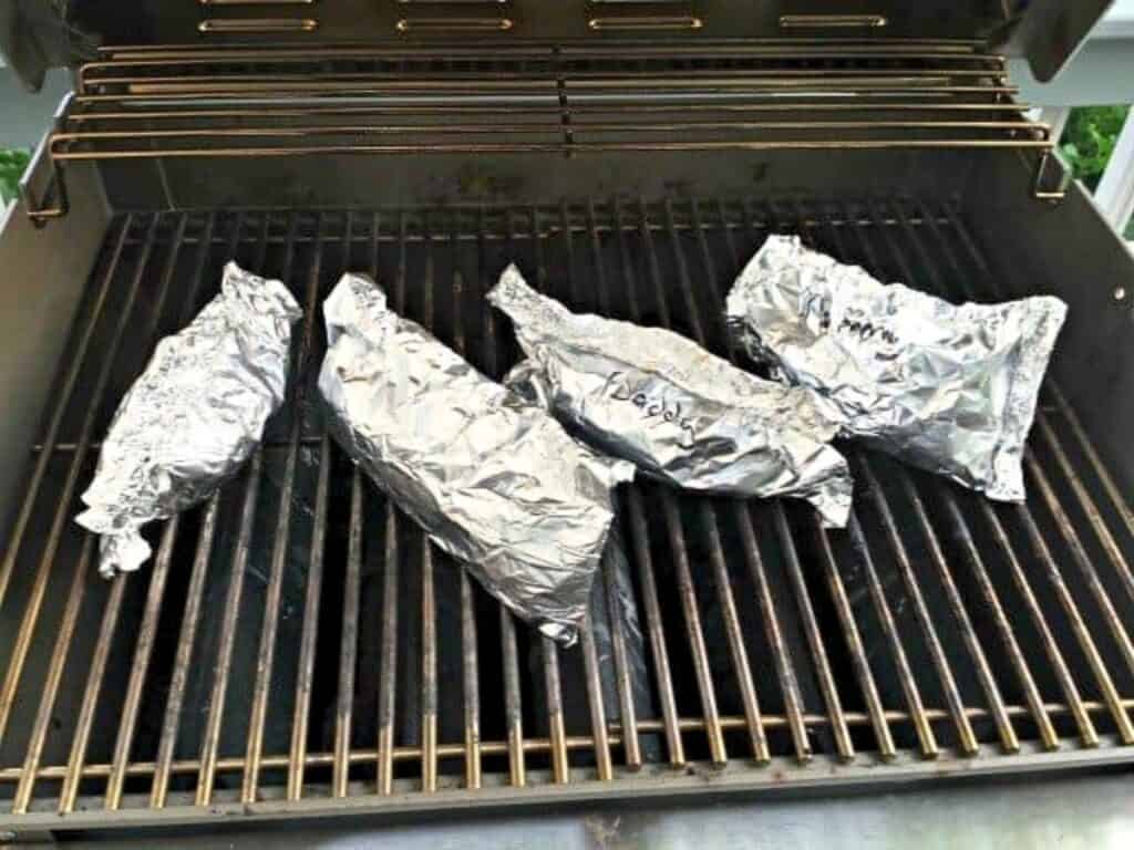 Four foil-wrapped food packages, including some labeled as grilled chicken pockets, are placed on the grates of an outdoor grill, with handwritten labels visible on some of the foil packets.