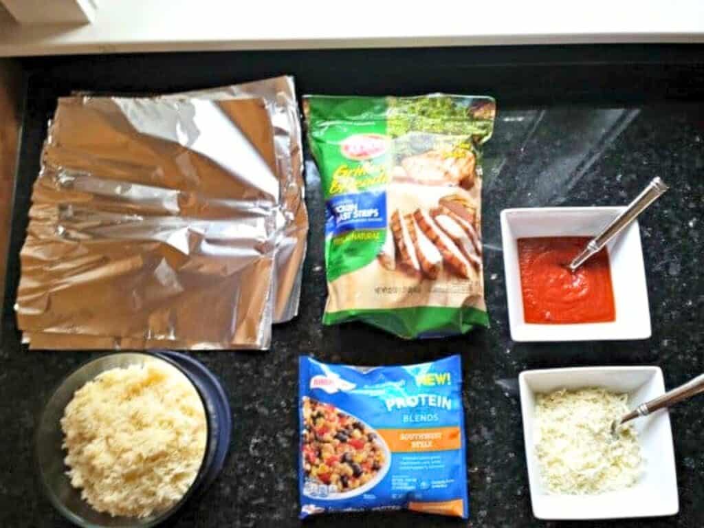 Foil sheets, shredded cheese, grilled chicken strips, mixed vegetables, tomato sauce, and grated parmesan cheese are arranged on a black countertop, ready to be assembled into delicious grilled chicken pockets.