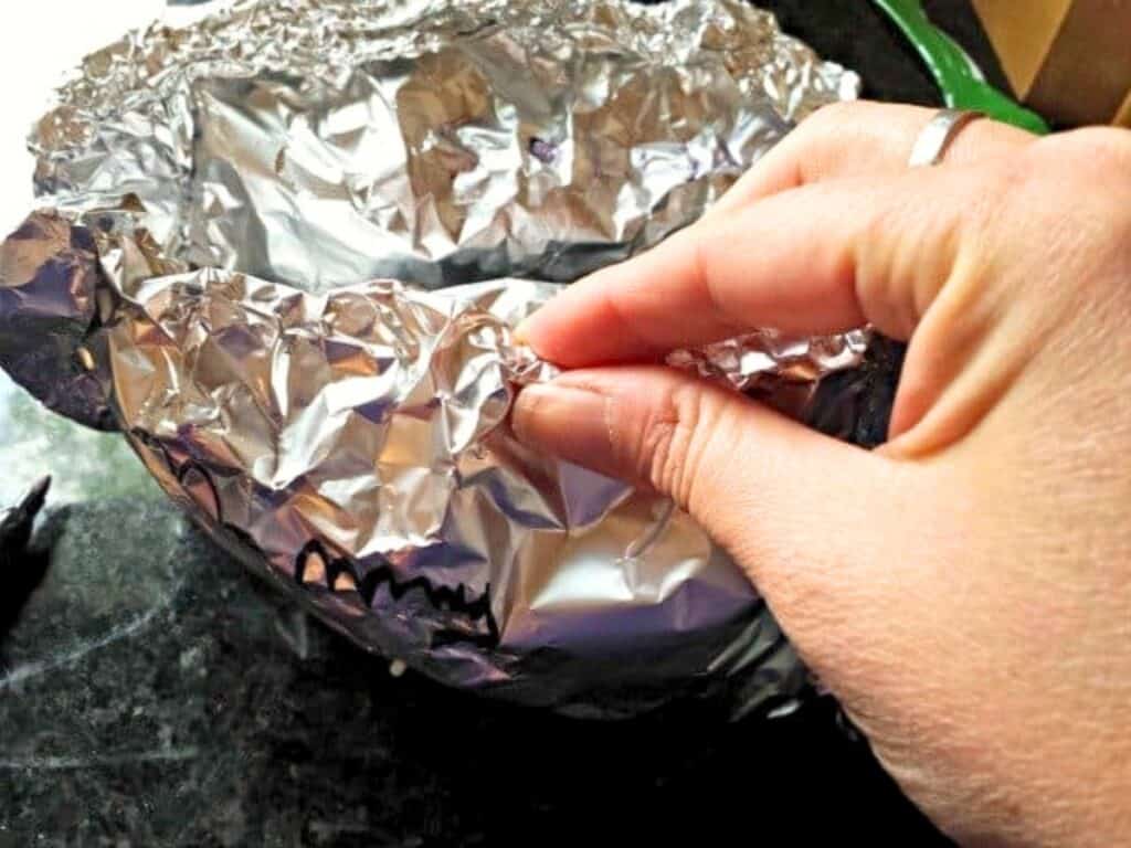 A hand presses aluminum foil around the rim of a bowl on a dark countertop, preparing to create perfect grilled chicken pockets.