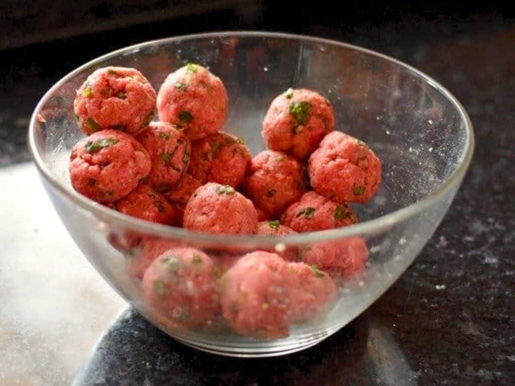 A glass bowl filled with raw meatballs mixed with herbs sits on a dark countertop.
