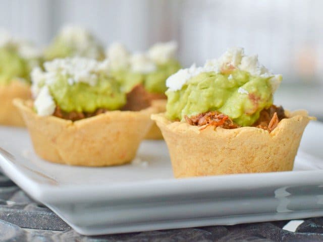 Image shows a close up head on view of Crockpot Mexican Beef in Masa Cups on a white tray, topped with guacamole and crumbled cotija cheese.