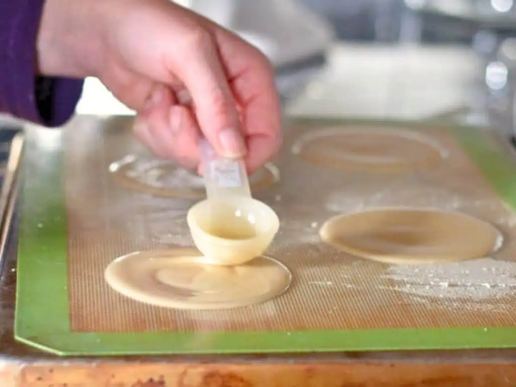 A hand pours batter from a measuring spoon onto a baking mat, forming round shapes for baking.