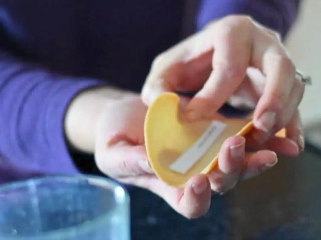 A person opens a fortune cookie and removes a paper fortune slip, with a glass of water visible in the foreground.