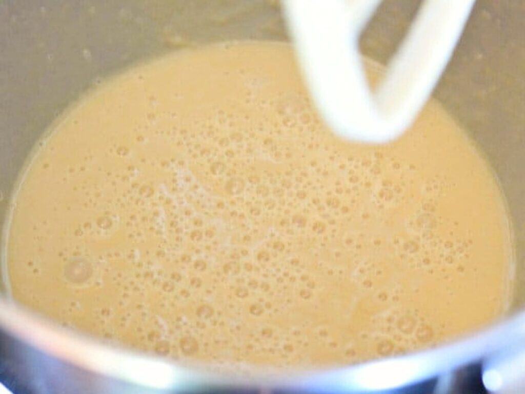 A mixing bowl filled with a light tan batter, with bubbles on the surface and a white mixing attachment partially visible above.