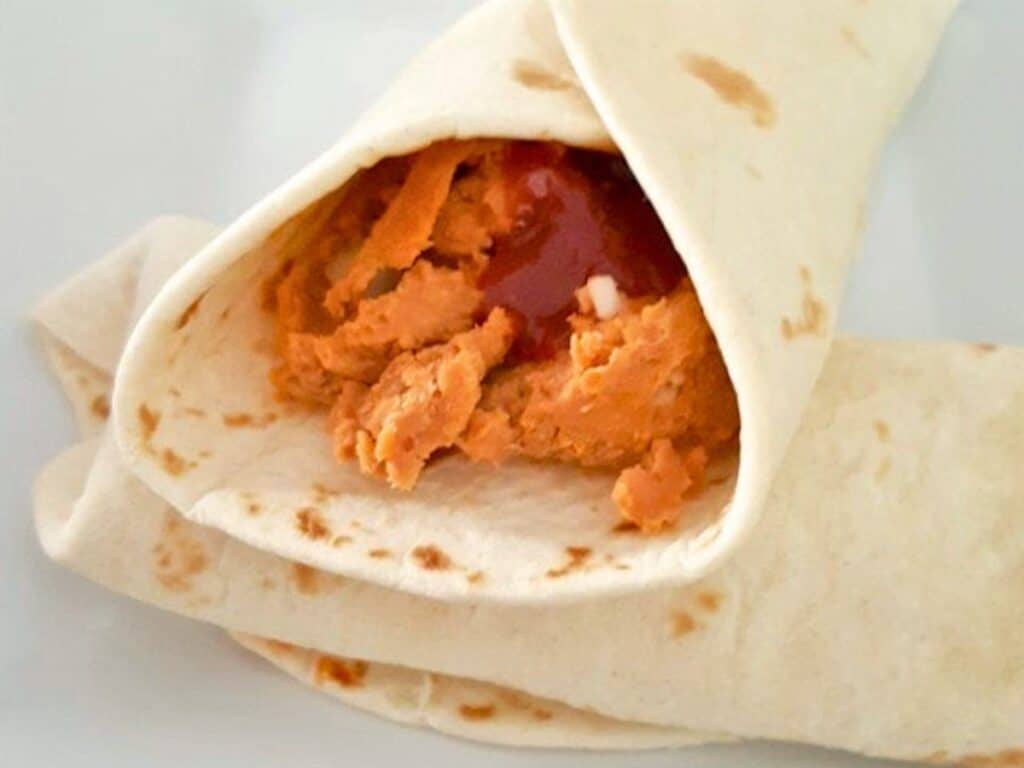 A flour tortilla is wrapped around a filling of homemade refried beans and a red sauce, partially open at one end, on a white surface.