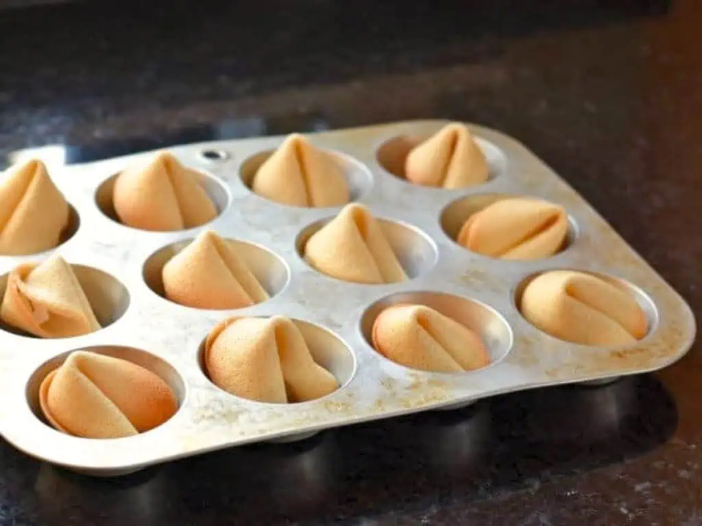 A muffin tin holds twelve golden-brown fortune cookies, each placed in an individual cup, on a dark countertop.