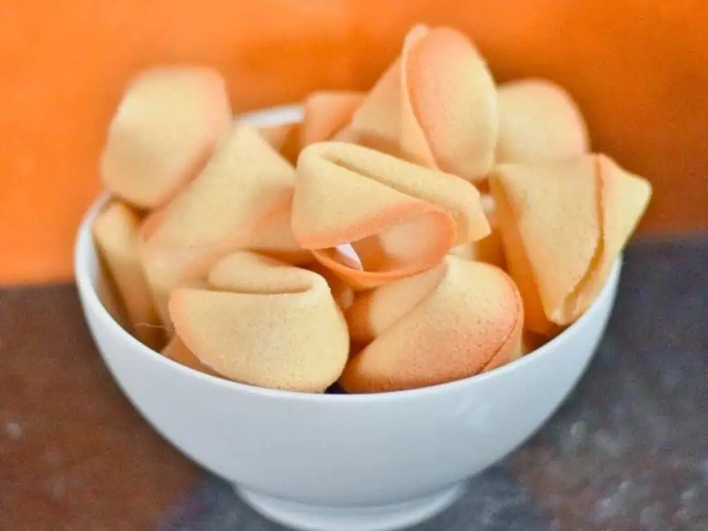 A white bowl filled with several fortune cookies is placed on a dark surface with an orange background.