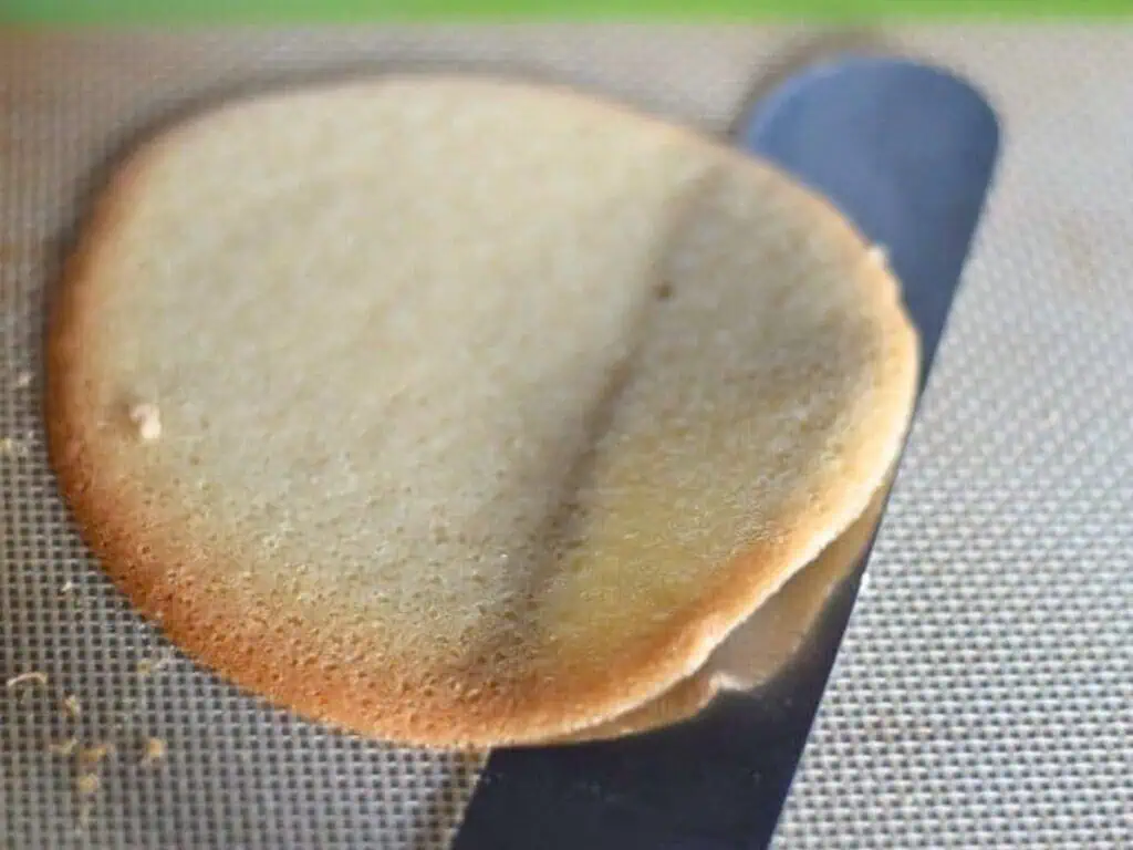 A thin, round baked cookie is being lifted from a silicone baking mat with a metal spatula.