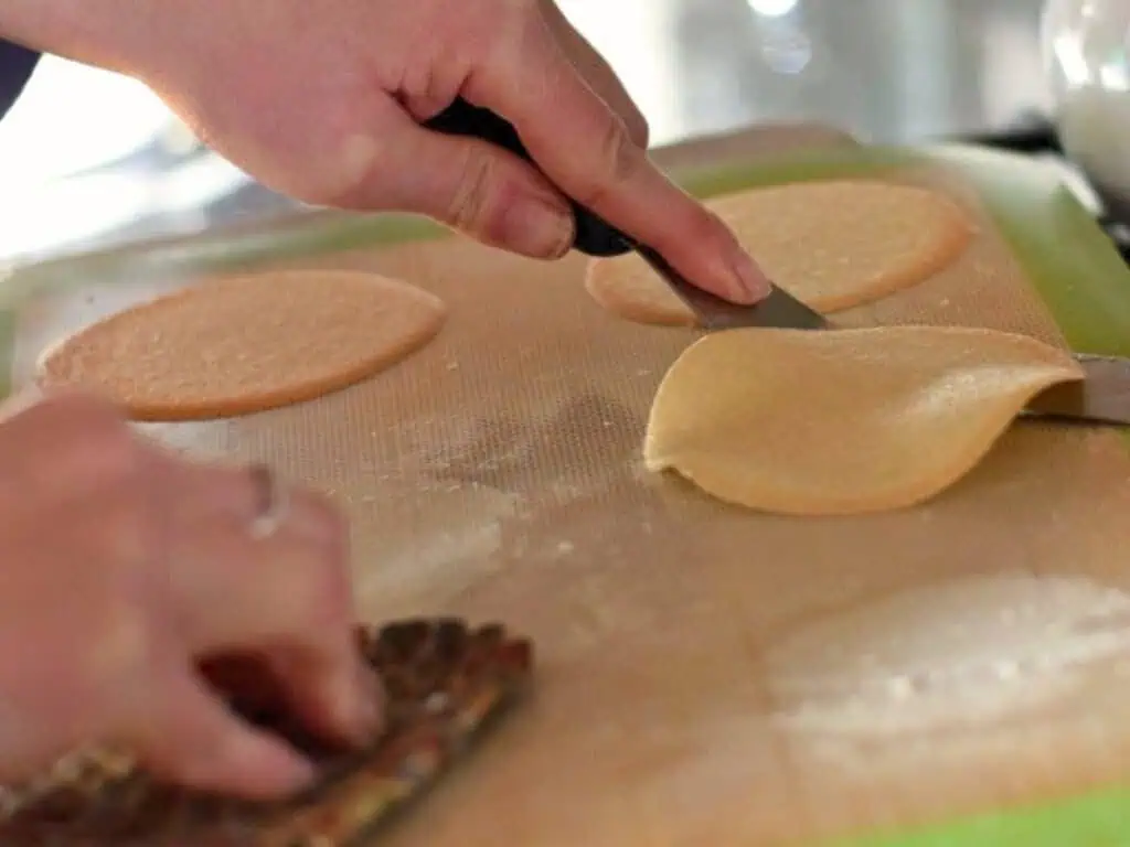 Hands are lifting a thin, round baked cookie off a silicone baking mat with a spatula; other cookies are on the mat.