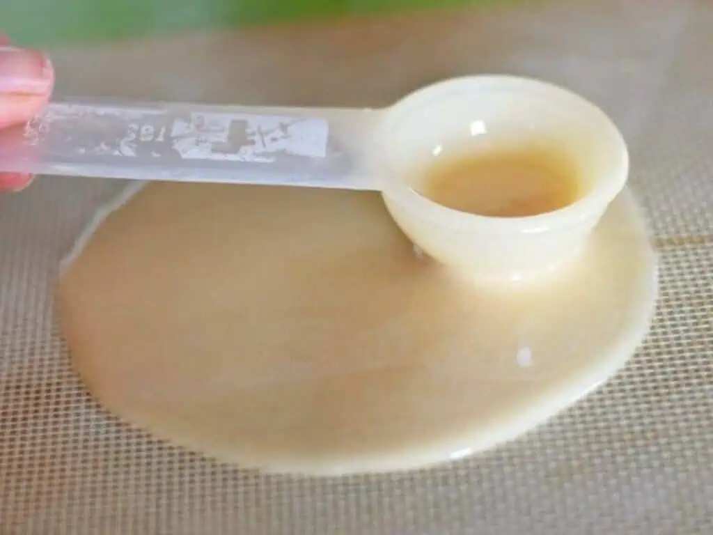 A plastic measuring spoon holding a thick, beige liquid, with some of the liquid spilled onto a textured surface below.