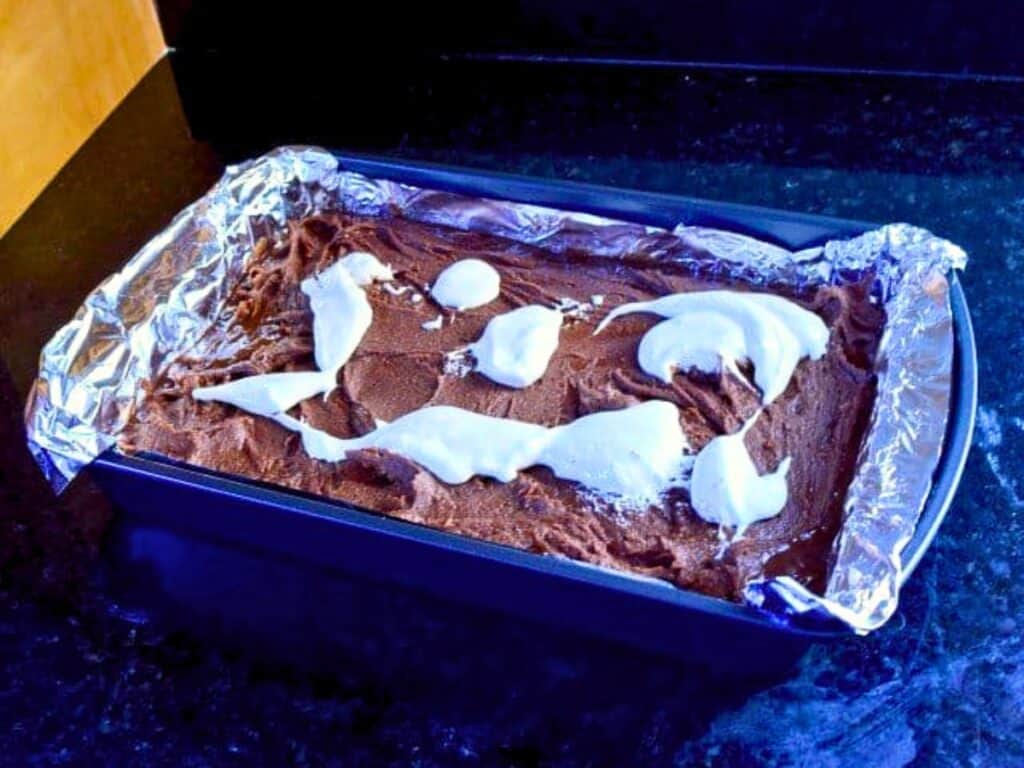 A metal loaf pan lined with foil holds rich chocolate s&rsquo;mores brownies batter, with irregular swirls of white cream or frosting spread on top, set on a dark countertop.
