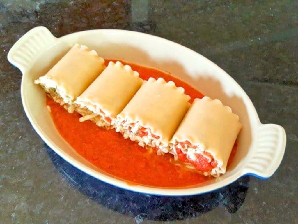 Four lasagna roll ups filled with cheese and tomato sauce are placed in a white oval baking dish with red sauce at the bottom, on a dark countertop.