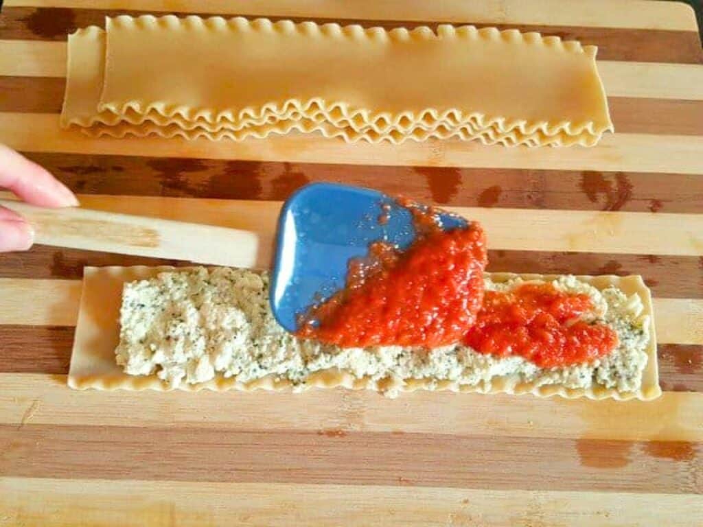 A spatula spreads tomato sauce over ricotta cheese on a flat lasagna noodle&mdash;perfect for preparing delicious lasagna roll ups&mdash;with another plain noodle above, all arranged on a wooden surface.