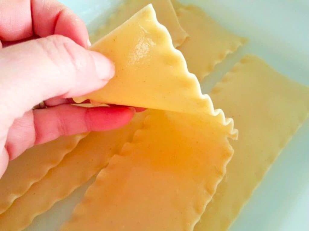 A hand holds a cooked lasagna noodle above a dish containing several other lasagna noodles, ready to be filled and rolled into delicious lasagna roll ups.