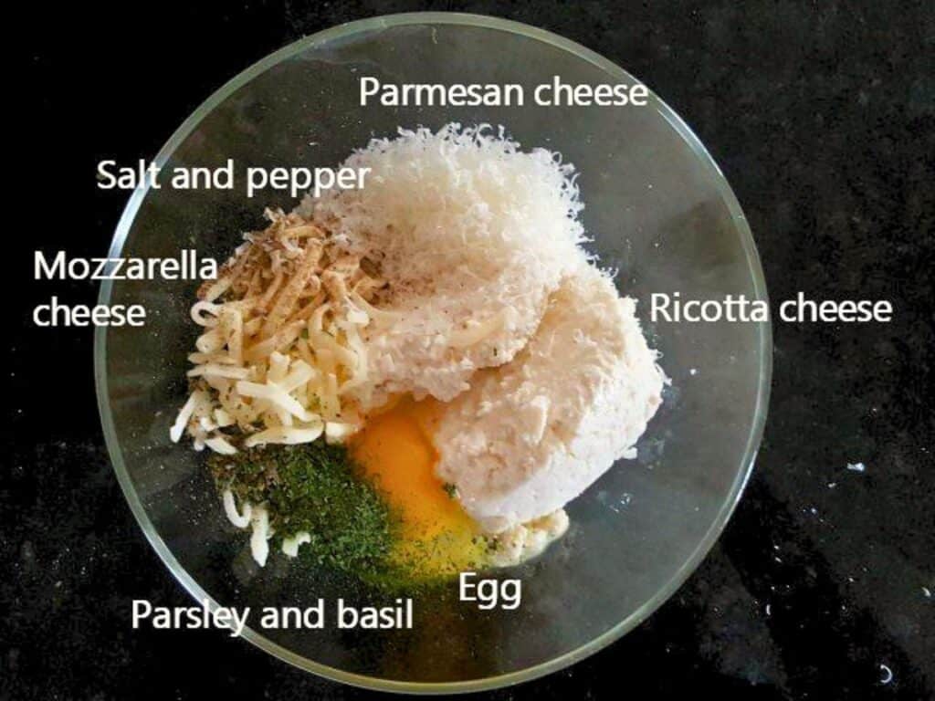 A glass bowl with ricotta, mozzarella, parmesan cheese, an egg, salt, pepper, parsley, and basil on a dark surface&mdash;perfect ingredients for classic lasagna roll ups.