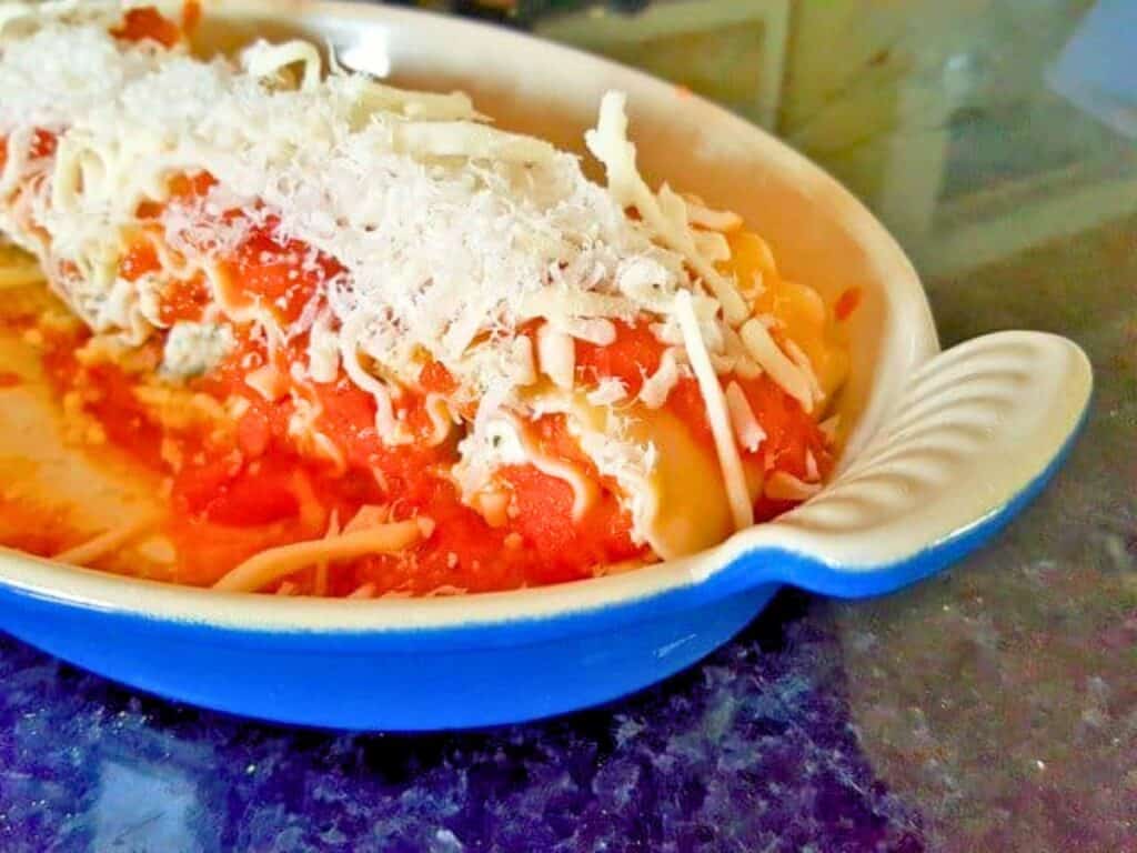 A baked pasta dish of lasagna roll ups in a blue oval dish, topped with tomato sauce and shredded cheese, sits on a dark countertop.