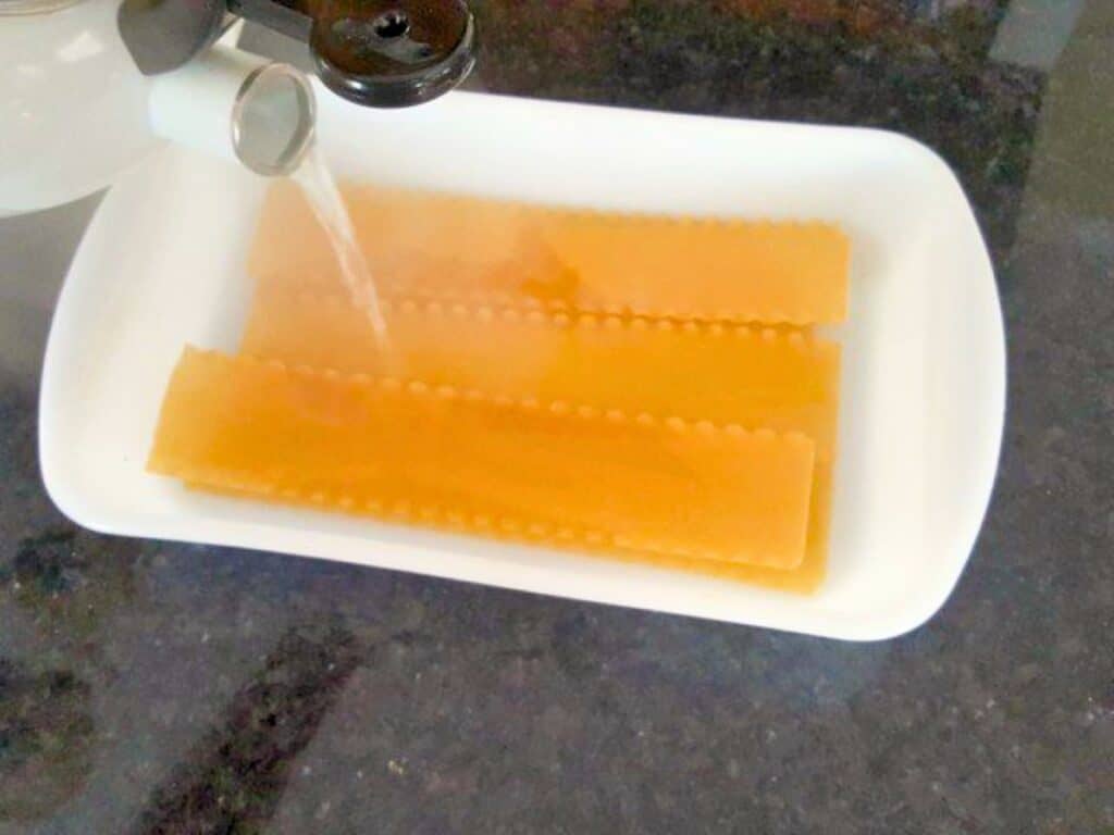 Three uncooked lasagna noodles in a white dish are being covered with hot water poured from a kettle, the first step in preparing delicious lasagna roll ups.
