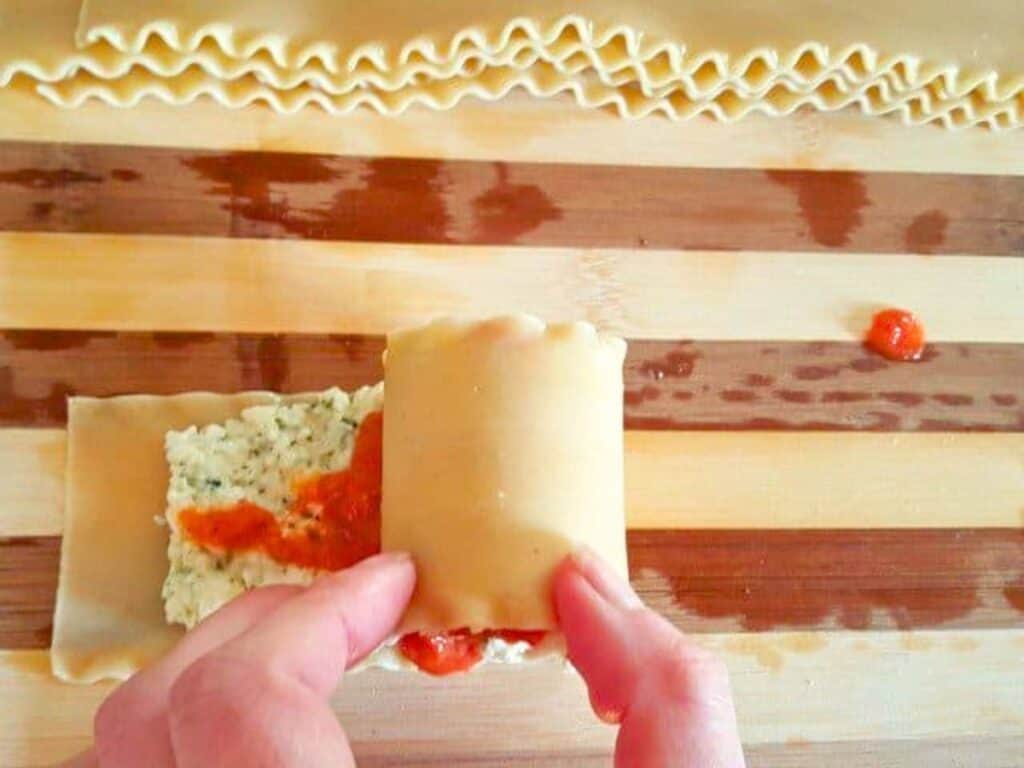 Hands rolling a lasagna noodle with ricotta filling and tomato sauce on a wooden surface; uncooked noodles are visible in the background, showcasing the delicious process of making homemade lasagna roll ups.