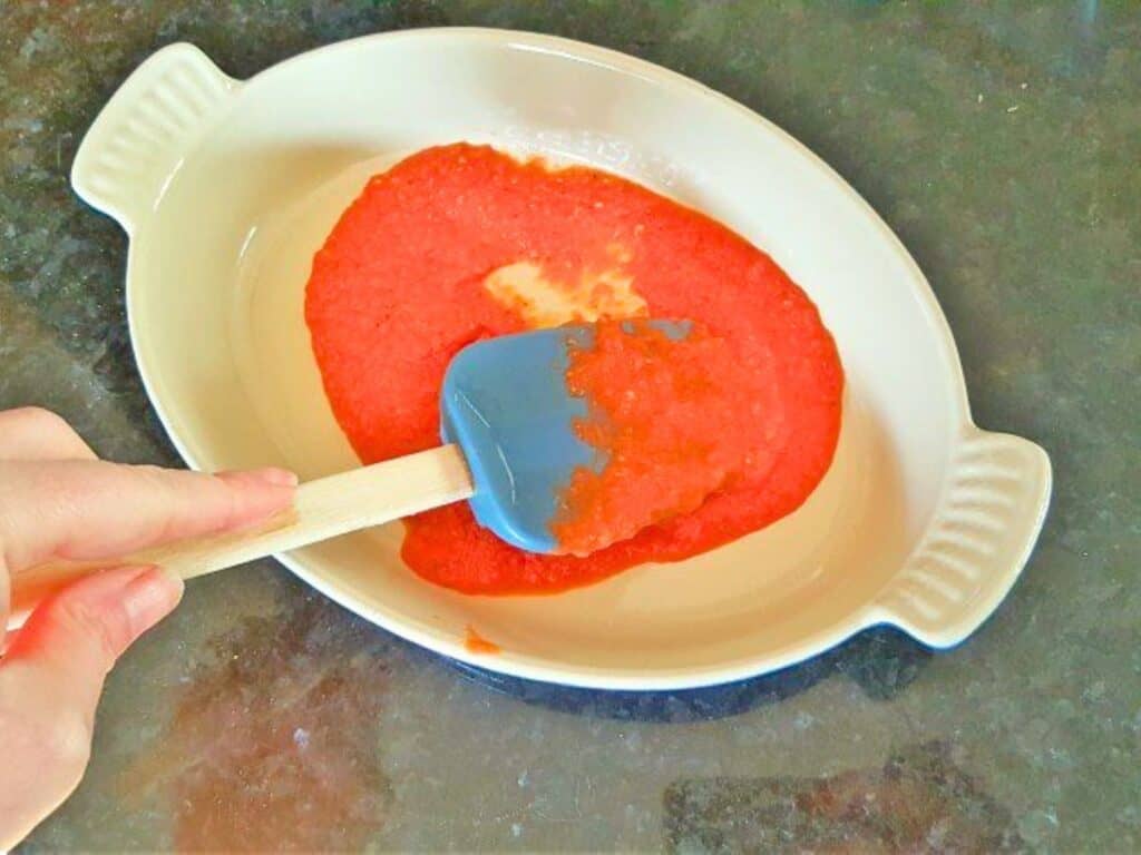A hand spreads red sauce in a white oval baking dish using a blue spatula, preparing the base for delicious lasagna roll ups.