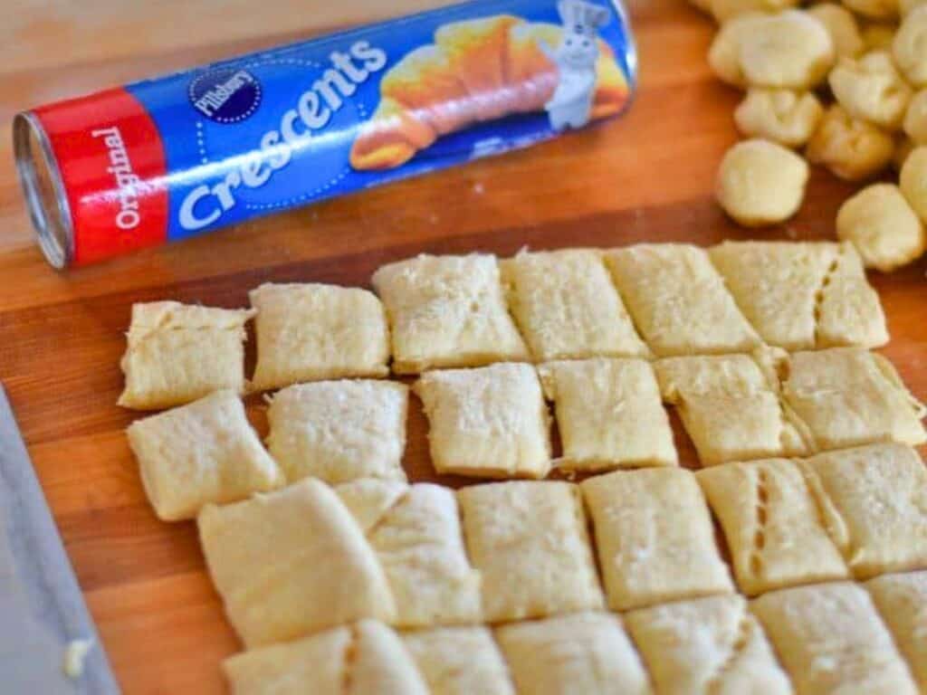 Squares of raw crescent roll dough are cut on a wooden board, with a can of Pillsbury Crescents in the background and small dough balls nearby.