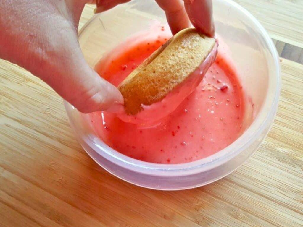 A hand dips a sponge cake into a bowl of pink strawberry sauce on a wooden surface, capturing the sweetness that inspires any great strawberry donuts recipe.