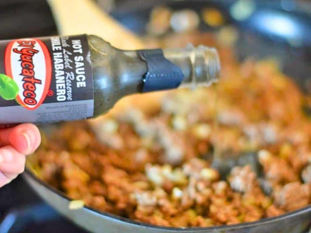 A hand pours El Yucateco hot sauce from a bottle onto browned ground meat cooking in a skillet.