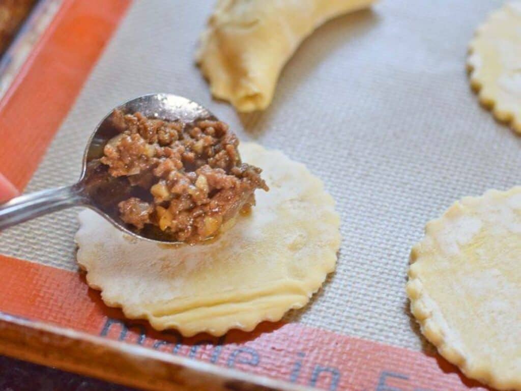 A spoon adds seasoned ground beef filling to a round piece of dough, with another dough circle and a folded pastry visible nearby on a baking mat.