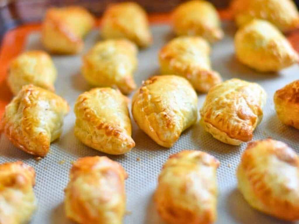 Golden-brown puff pastry turnovers arranged in rows on a baking sheet lined with a silicone mat.