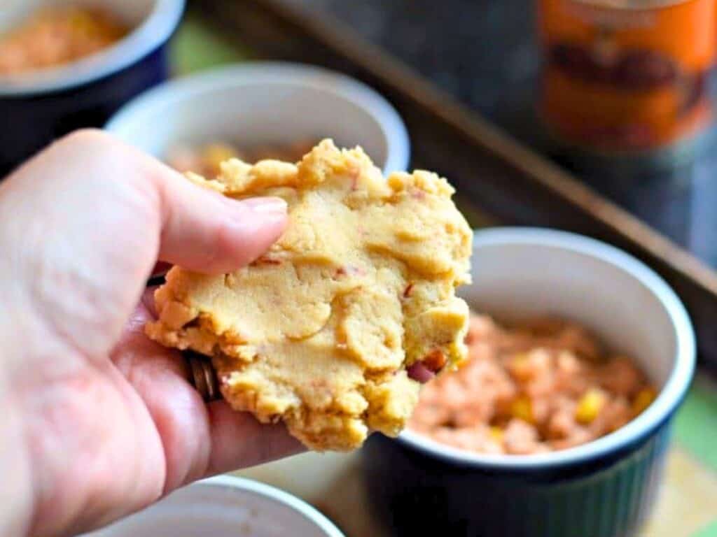 A hand holds a round piece of uncooked dough over a ramekin filled with a chunky mixture for tamale pot pies; other filled ramekins are visible in the background.
