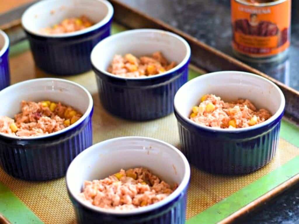 Six blue ramekins filled with a chunky mixture, possibly tuna and corn, are placed on a baking sheet with a silicone mat&mdash;perfect individual servings reminiscent of tamale pot pies. A can is visible in the background.