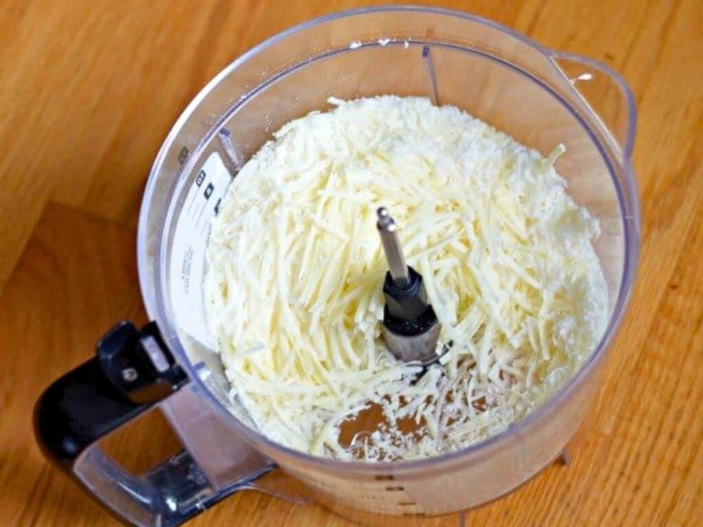 A food processor bowl filled with shredded cheese, perfect for making cacio e pepe, is placed on a wooden surface.