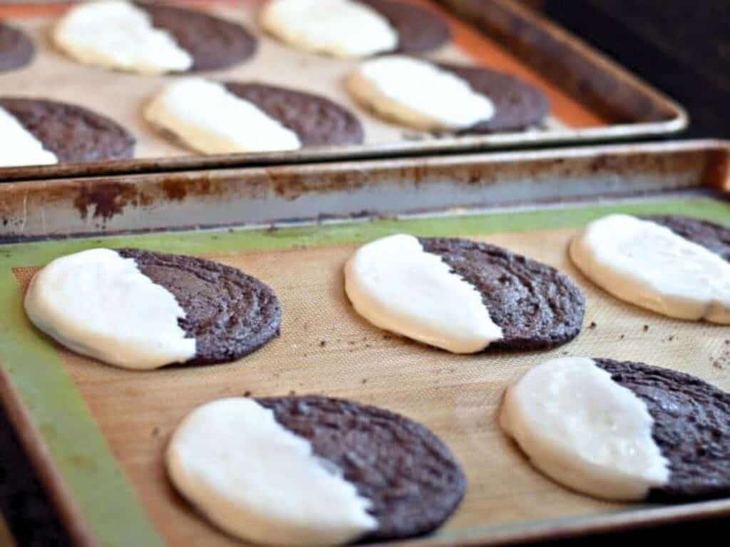 Chocolate cookies on baking sheets, each half coated in white icing, are cooling on silicone mats.