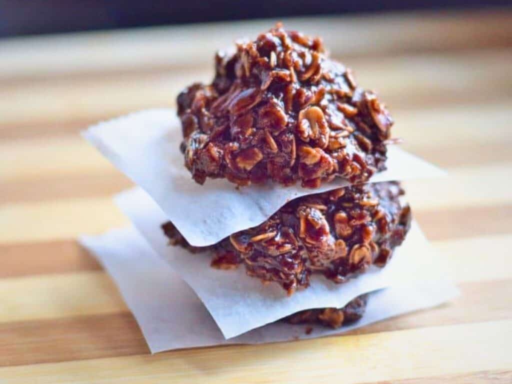 Three Nutella no bake cookies are stacked on parchment paper squares on a wooden surface, showcasing their rich chocolate oatmeal layers.