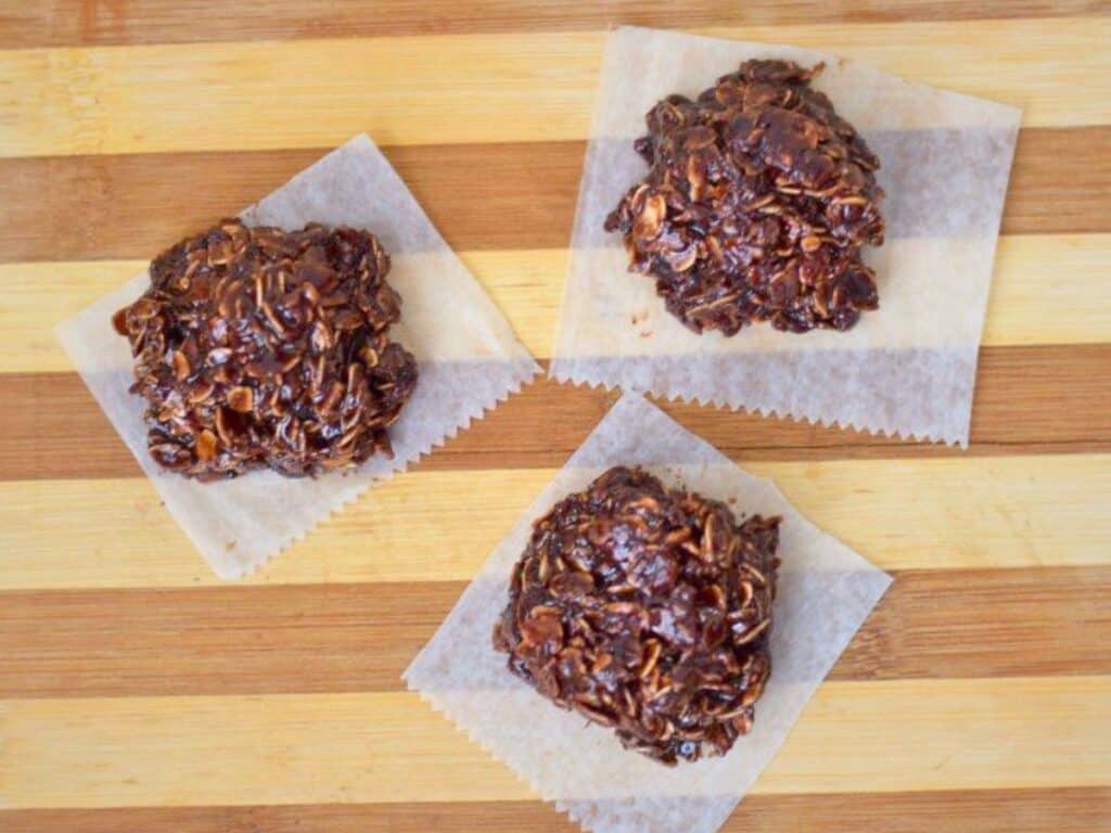Three chocolate oatmeal Nutella no bake cookies sit on wax paper squares atop a striped wooden surface.