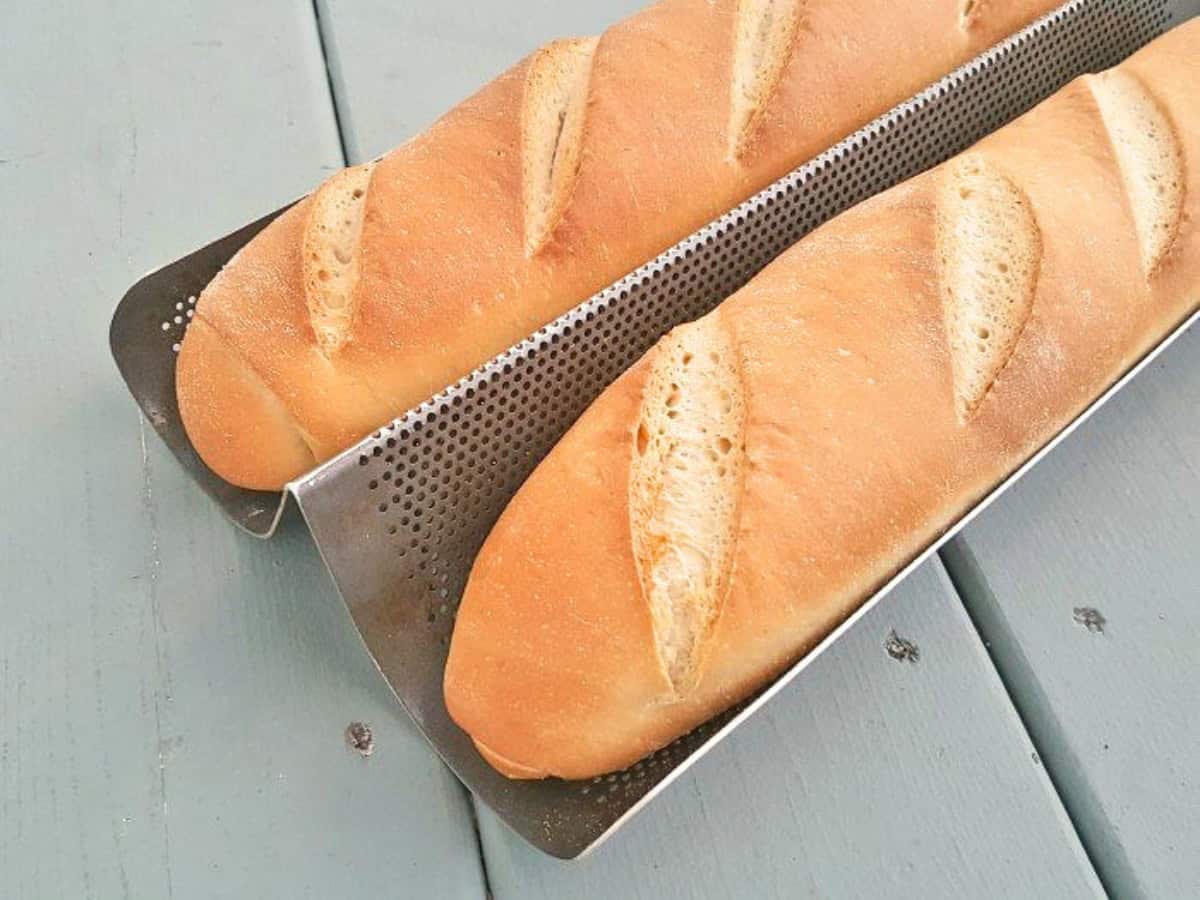 Two freshly baked baguettes with slits on top, made from a classic baguette recipe, are resting on a perforated metal baking tray placed on a light blue wooden surface.