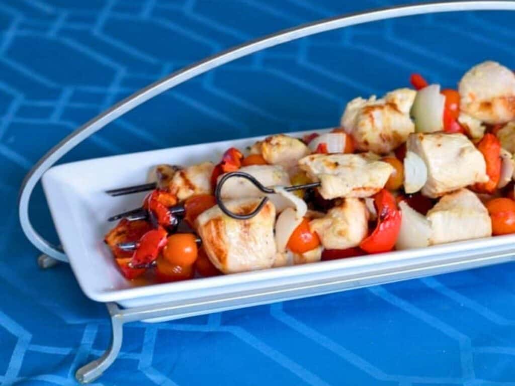 Lemon marinated chicken kebabs and fresh vegetable skewers are arranged on a rectangular white plate with a metal handle, set on a blue patterned surface.