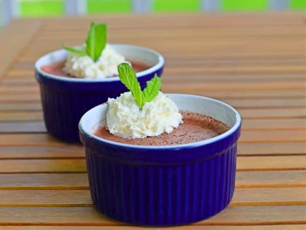 Two blue ramekins filled with cherry mousse, each topped with whipped cream and a mint leaf, placed on a wooden table.