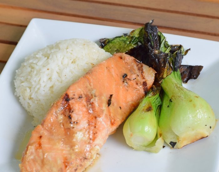 Image shows a Bite of miso glazed salmon with rice on a fork with the rest of the plate in the background.