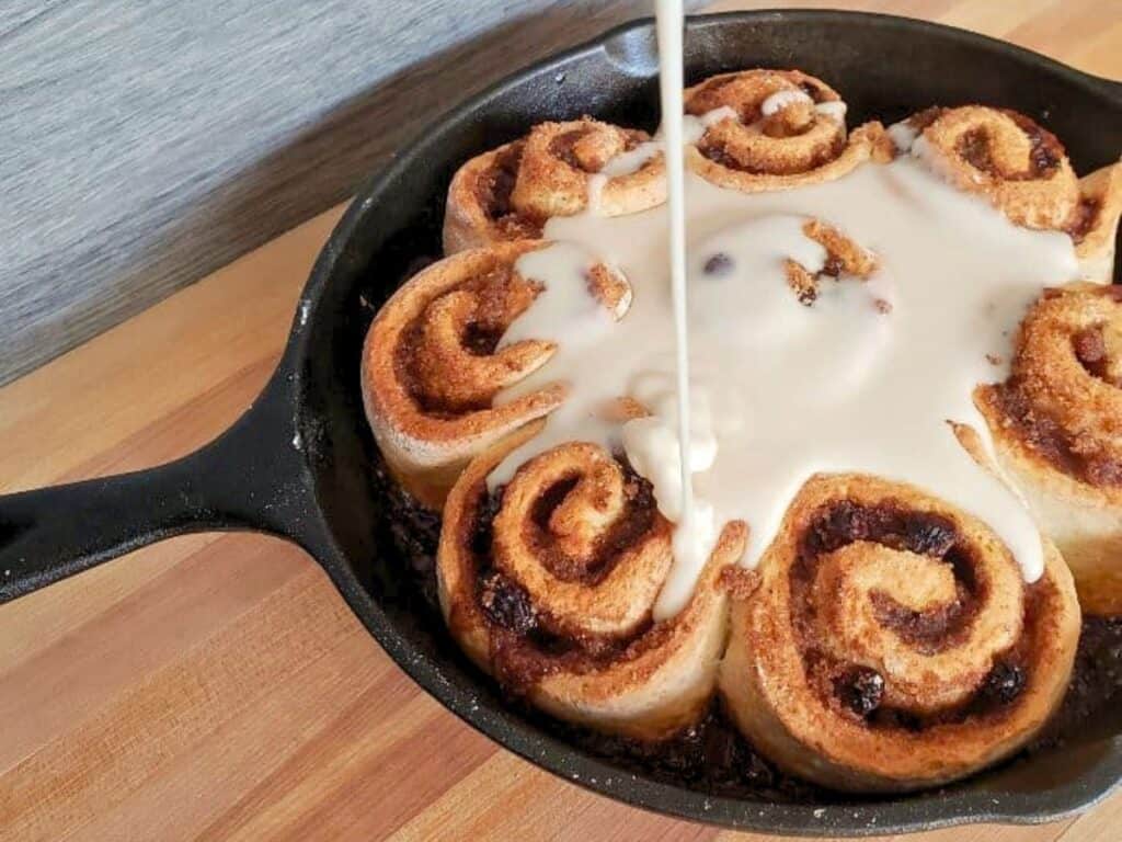 A skillet of no yeast cinnamon rolls sits on a wooden surface, with icing being poured over the rolls from above.