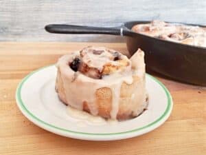 A no yeast cinnamon roll topped with icing sits on a white plate, with a pan containing more rolls in the background on a wooden surface.