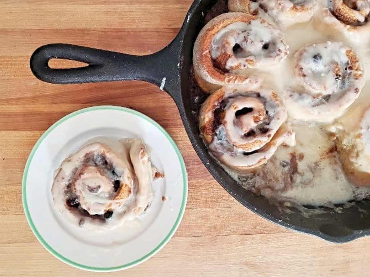 A cast iron skillet with no yeast cinnamon rolls sits on a wooden surface. One cinnamon roll has been served on a small white plate with a green rim.