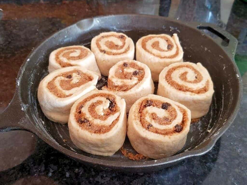 Unbaked no yeast cinnamon rolls with raisins arranged in a cast iron skillet on a dark countertop.