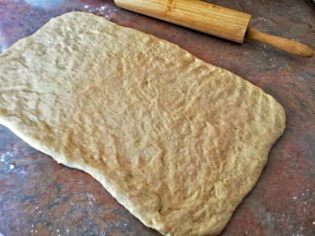 A rectangular sheet of dough for no yeast cinnamon rolls is rolled out on a floured surface with a wooden rolling pin nearby.